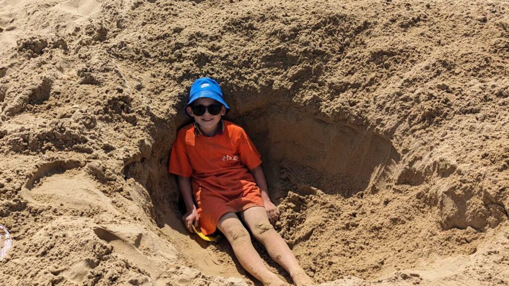 ID: a Branch Up young person is in a hole dug into the sand at the beach, smiling and wearing sunglasses.